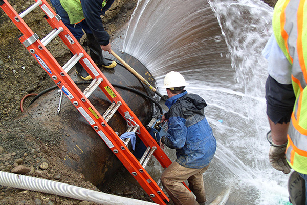 A worker climbing down a ladder to make a water pipe repair, while water gushes out.