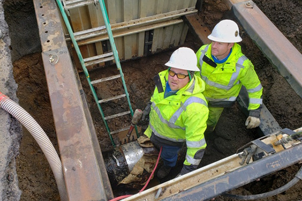 two workers with yellow green jackets and hard hats in a trench