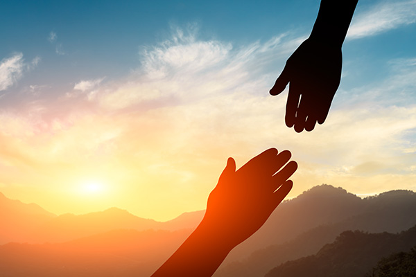 A close up of a hand reaching out to help another, with a sunset behind them.