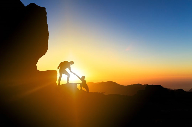 A person helping another person up a mountain trail with a sunrise behind them.