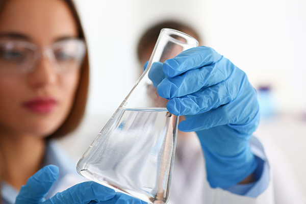 A woman wearing blue gloves and eye protection, holds a beaker of crisp water and examines it.